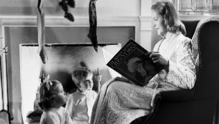 Black and white photo of a woman reading to kids in front of a fireplace.