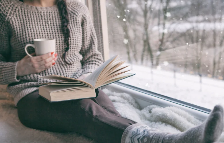 A person sitting by a window reading a book. It's snowing outside.