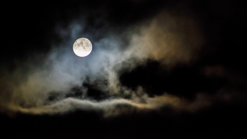 Photo of a dark and cloudy night sky with a brilliant white full moon peeking through the clouds