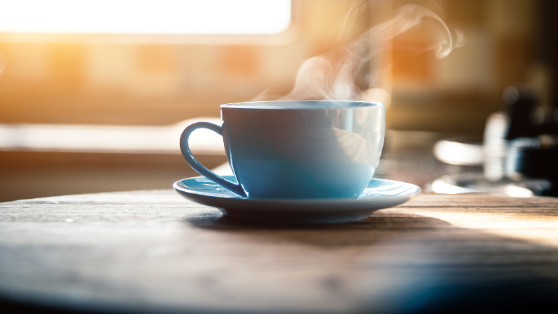 Photo of a table in front of a window, with a blue cup of steaming coffee or tea.